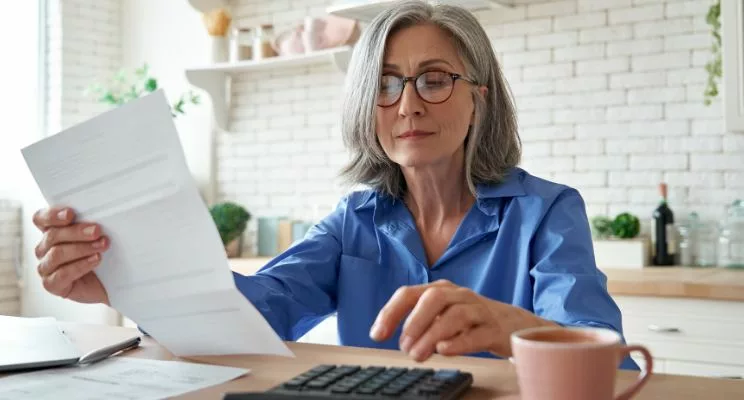 Older woman in kitchen staring calmly at paper in her hand with calculator and coffee mug