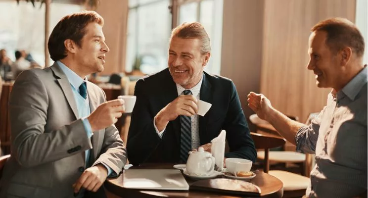 three businessmen sit in a cafe, smiling and talking while drinking coffee